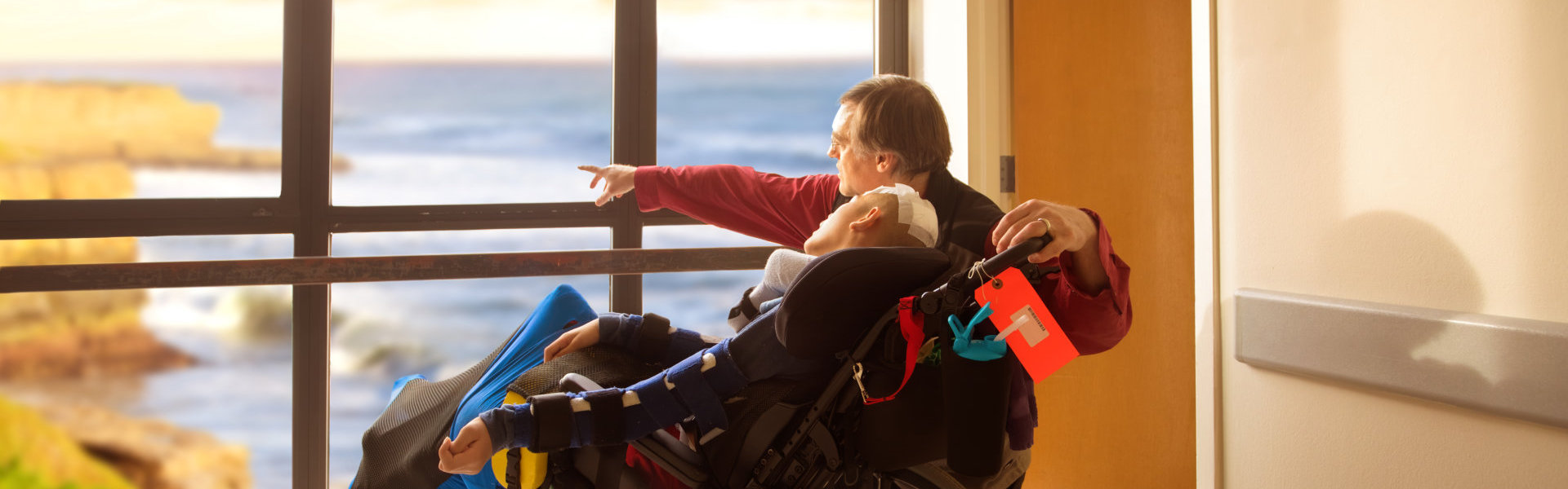 father kneeling next to his son in wheelchair looking out hospital window