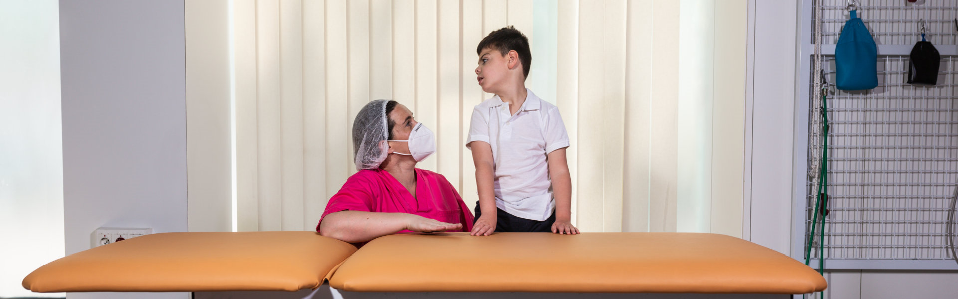 woman in a pink gown, mask and protective cap observes and gestures with one hand at a child with multiple disabilities while helping him with rehabilitation to learn to stand up
